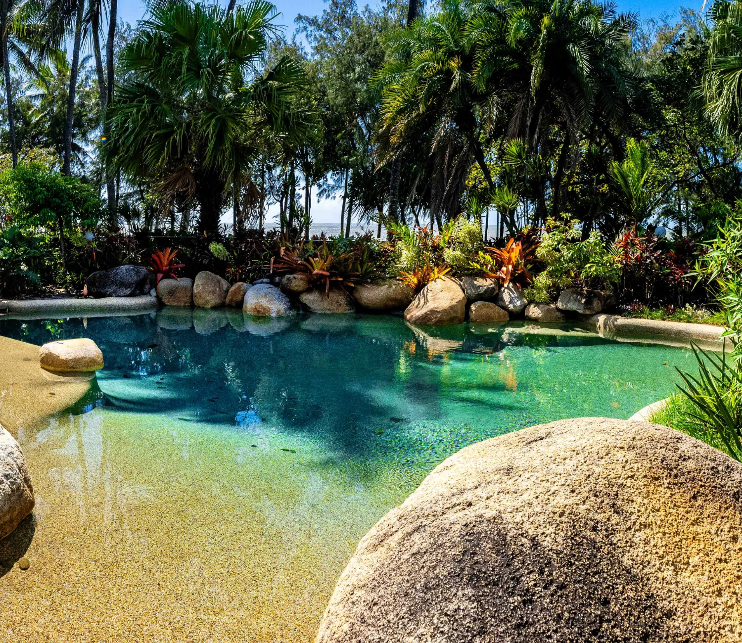 Melaleuca Resort Pool showing the rocks and gardens and behind the beautiful palm trees, you can see the beach.